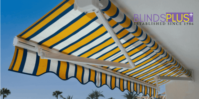 Yellow, blue, and white striped retractable awning installed on a modern home exterior with BlindsPlus logo displayed in the top corner.