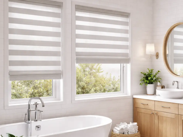 Modern bathroom interior with dual zebra roller blinds on white-framed windows, freestanding bathtub, wooden vanity, round mirror, and indoor plant in natural light.